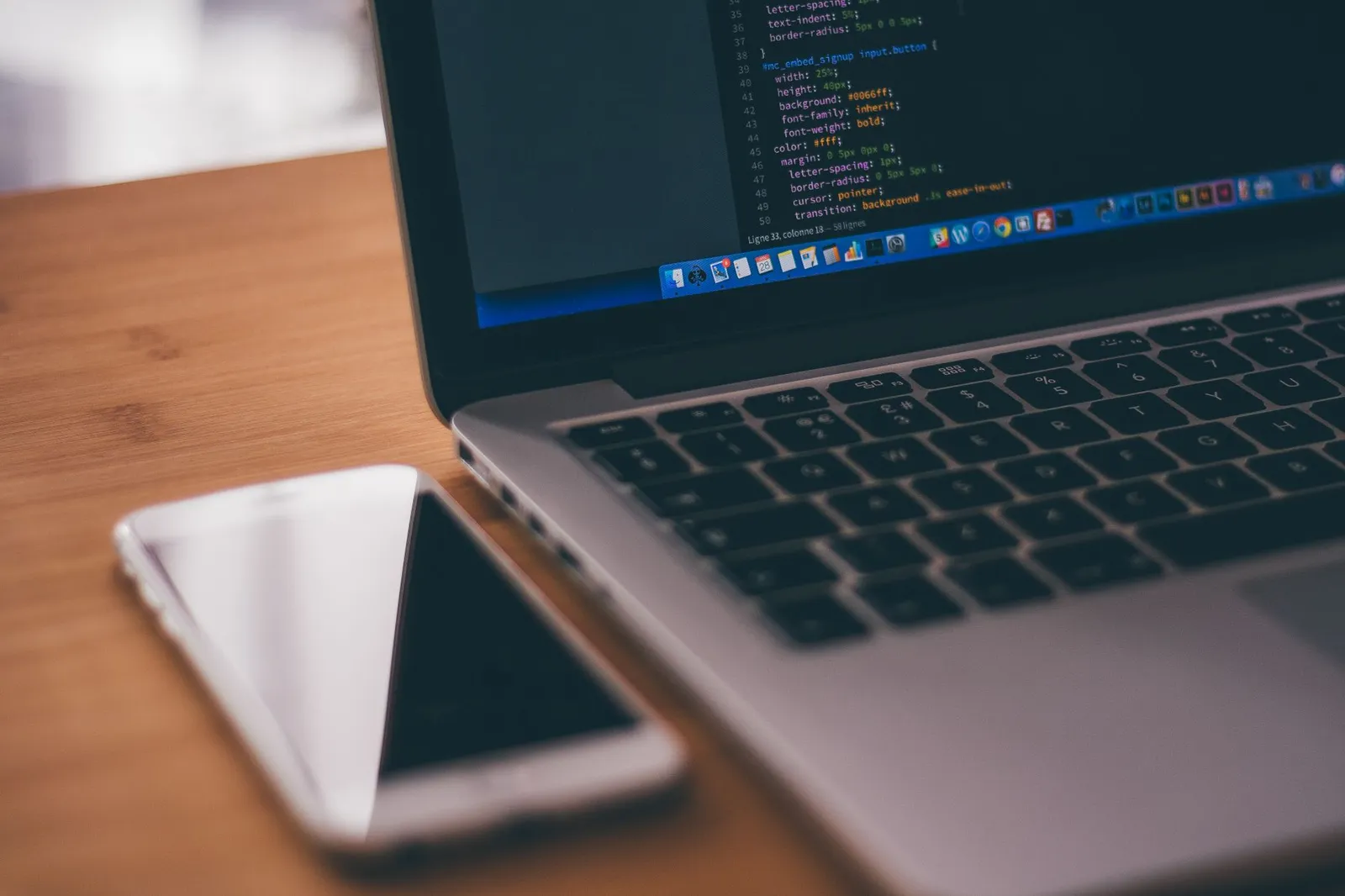 Laptop showing code next to a smartphone on a desk — an app being built and tested on the phone it will run on
