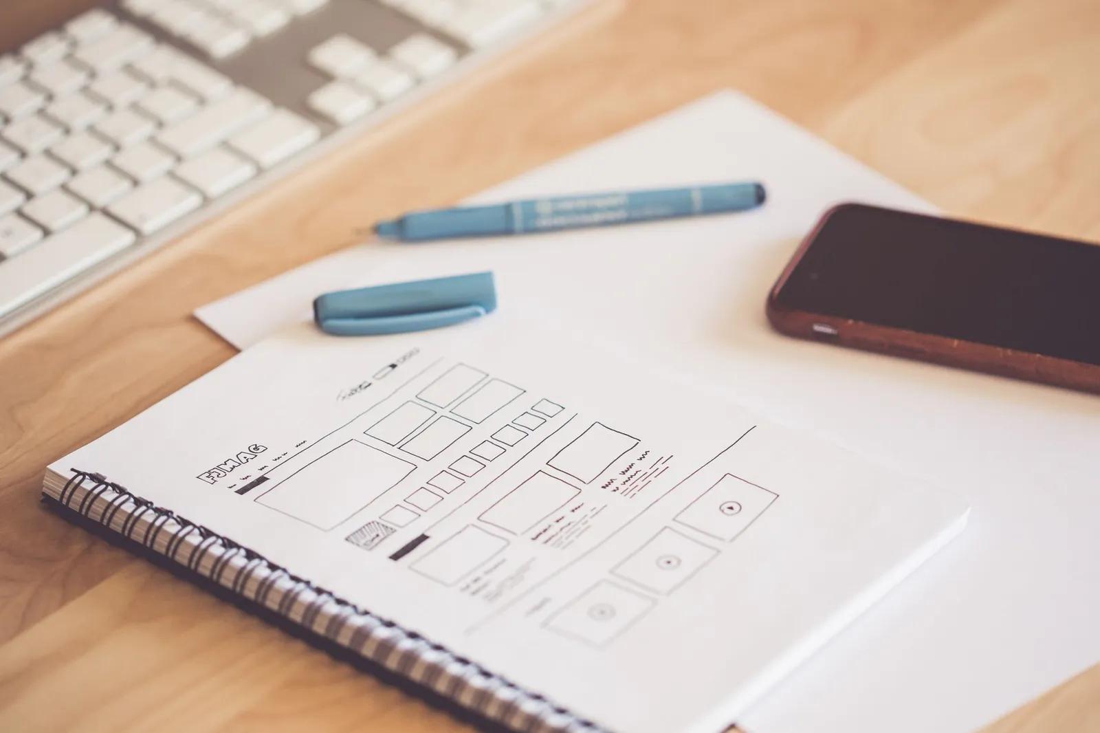 Sketchbook of app screen wireframes beside a phone and keyboard — screens are planned on paper before any code