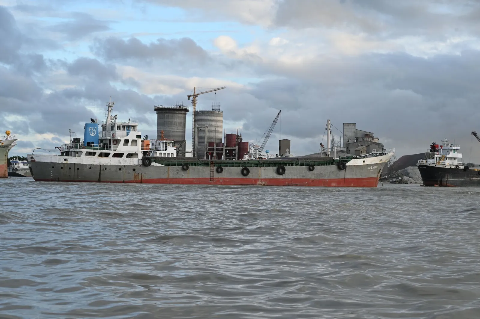 Cargo ship on the water at Chattogram Port, with cranes and storage silos behind — the shipping trade many Chattogram businesses serve