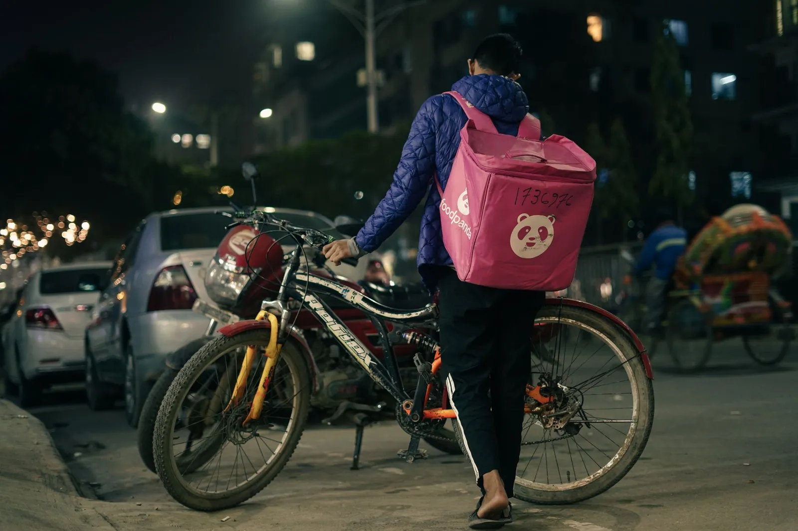 Delivery rider with a food bag walking a bicycle along a city street at night — the kind of order an app sends out