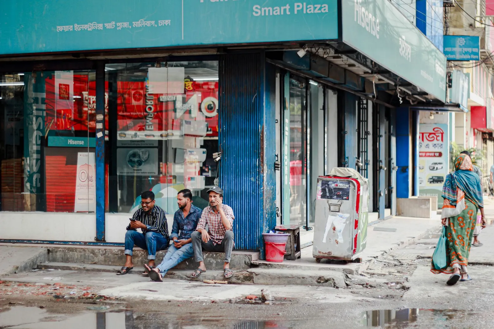 Men sitting outside an electronics shop on a Dhaka street — the kind of local business that still has no website