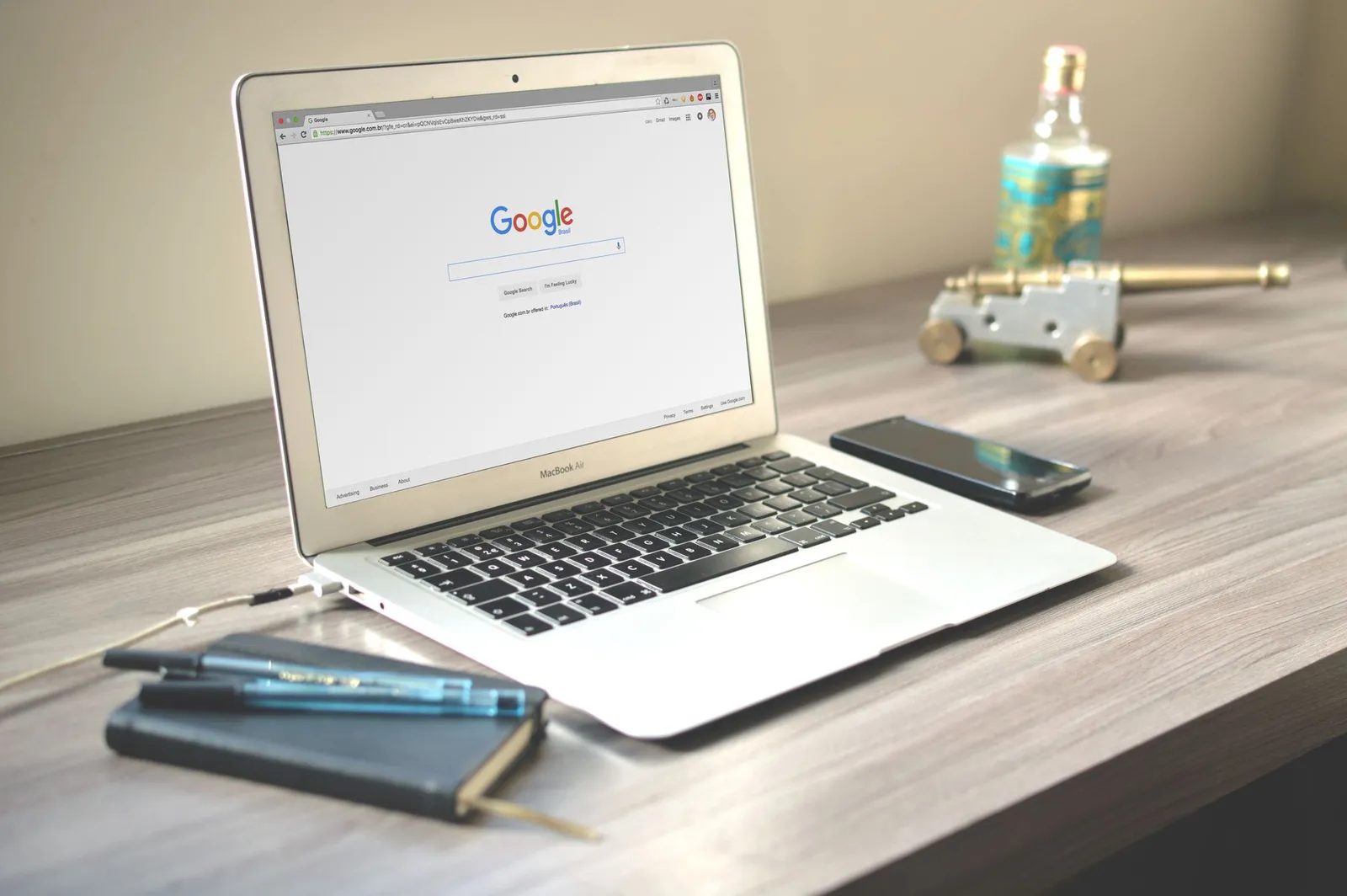 Laptop on a wooden desk open on the Google search page, next to a phone and notebook — where SEO work is checked