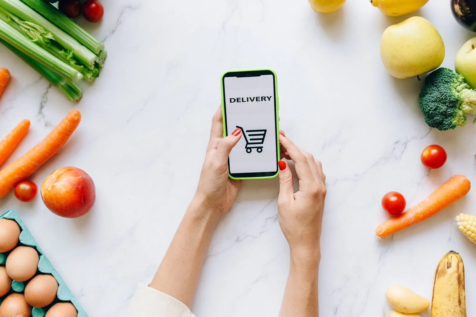 Hands holding a phone with a delivery shopping app open, surrounded by fresh vegetables and fruit