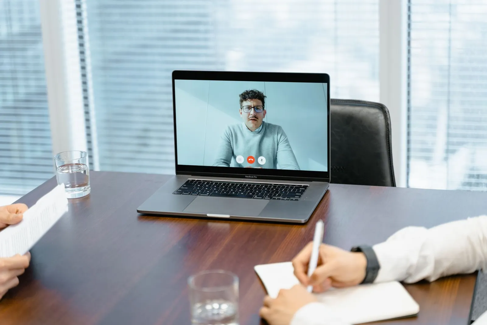 Laptop on a meeting-room table showing a video call while two people take notes, the way a remote website project is reviewed