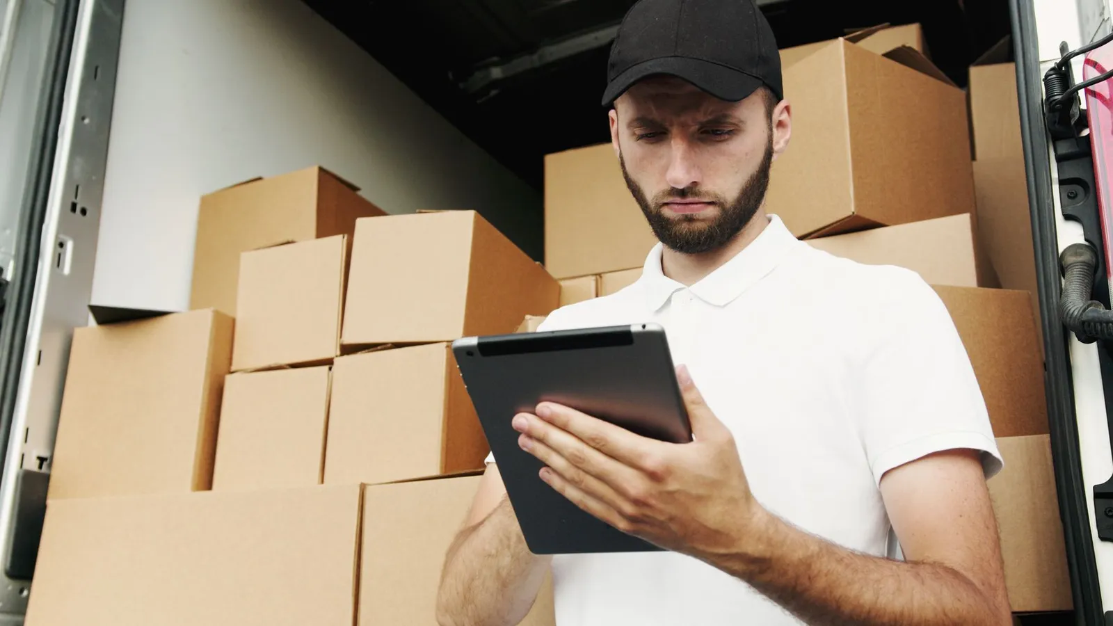 Delivery worker checking shipments on a tablet beside stacked boxes — logistics data an app reads from the same back end as the website